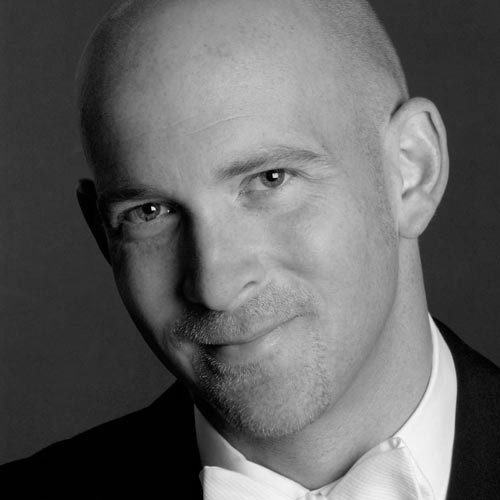 A bald man wearing a suit jacket and shirt with white bow tie.