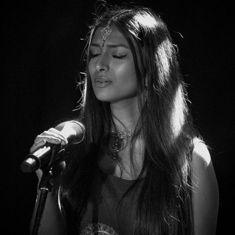 A woman singing into a microphone. She has long dark hair and is wearing jewellery around her neck and on her forehead.